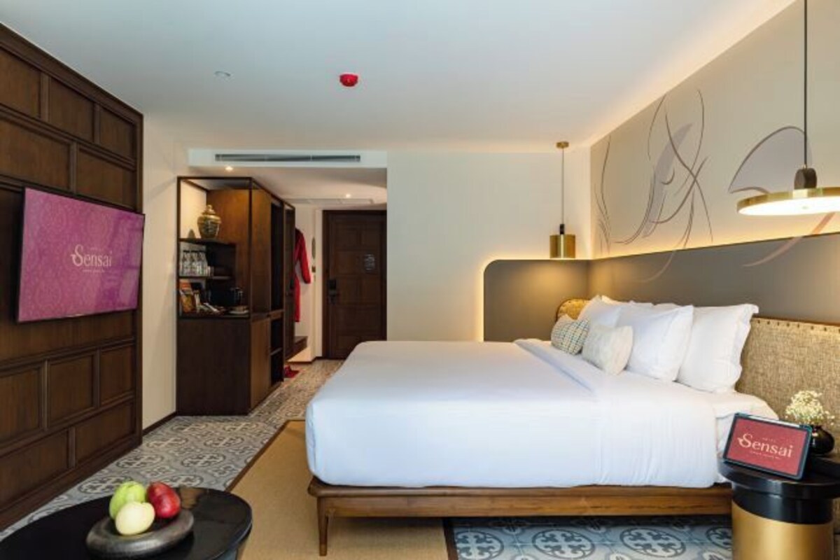 Hotelkamer met een bed, een televisie, een nachtkastje met een fruitschaal, en een bord met de naam van het hotel.