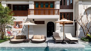 Un espace piscine avec des chaises longues et un canapé en osier, un vélo, et un bâtiment doté de balcons et de volets en bois.