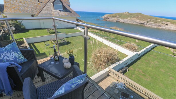 Eine Terrasse mit Korbmöbeln, einem Tisch mit Flasche und Gläsern sowie Blick auf Meer und Klippen.
