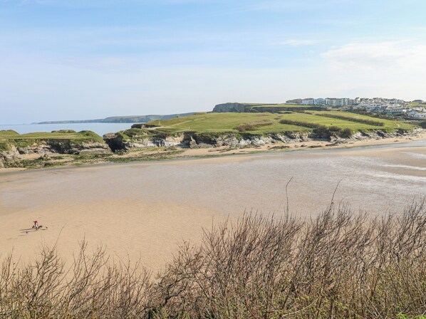 A coastal beach with a sandy shore, grassy cliffs, and a distant view of buildings. - Island View (Newquay)