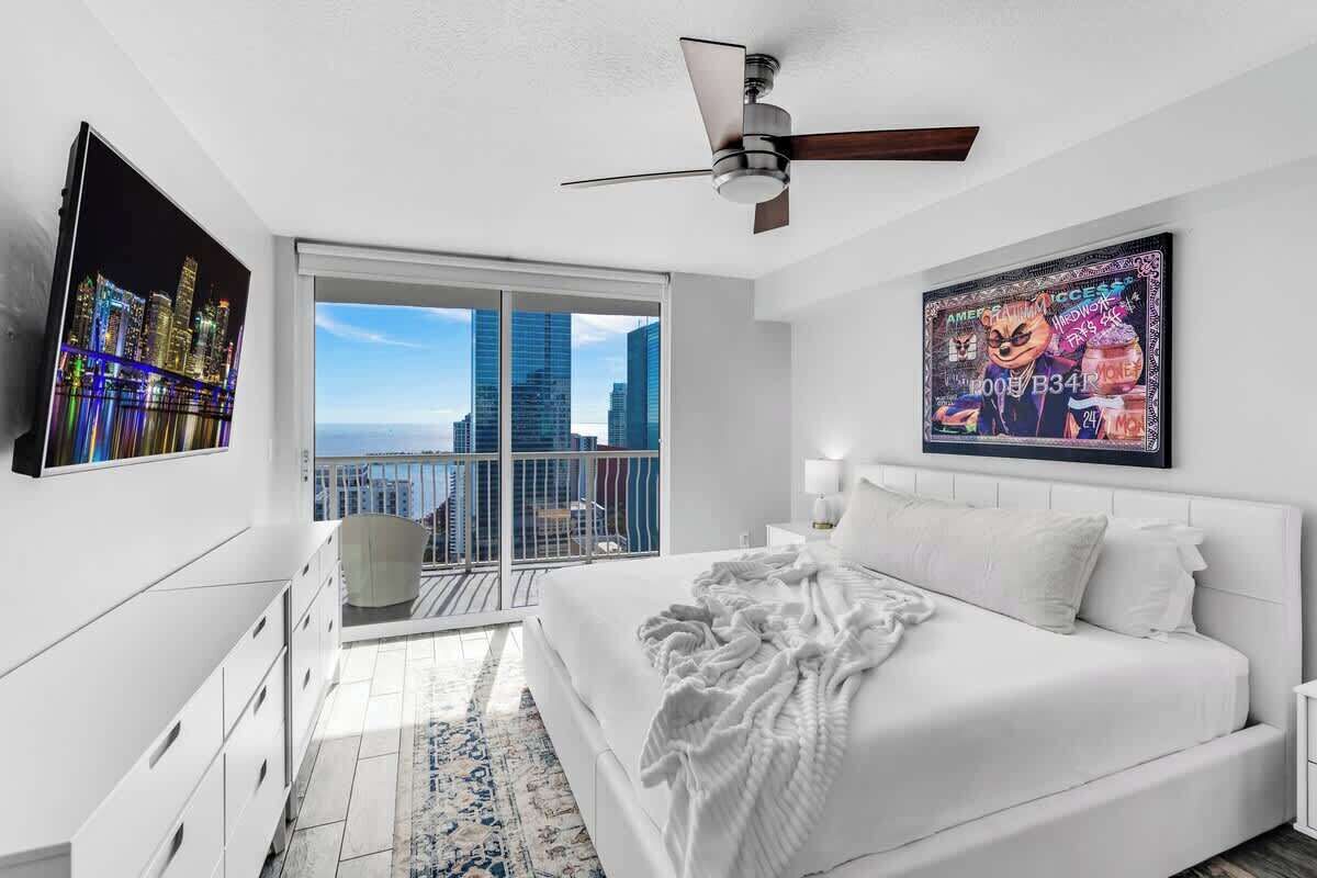 Quarto moderno com uma cama grande, ventilador de teto, varanda com vista para a cidade e uma obra de arte emoldurada na parede.