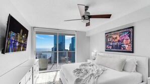 Quarto moderno com uma cama grande, ventilador de teto, varanda com vista para a cidade e uma obra de arte emoldurada na parede.