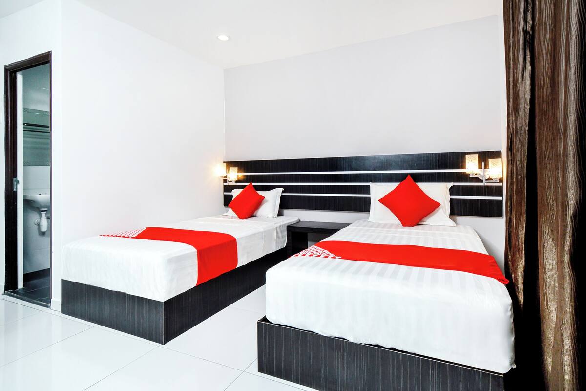 Een moderne hotelkamer met twee bedden, elk met witte beddengoed en rode accenten, een zwart-wit hoofdeinde en een klein nachtkastje met een lamp.