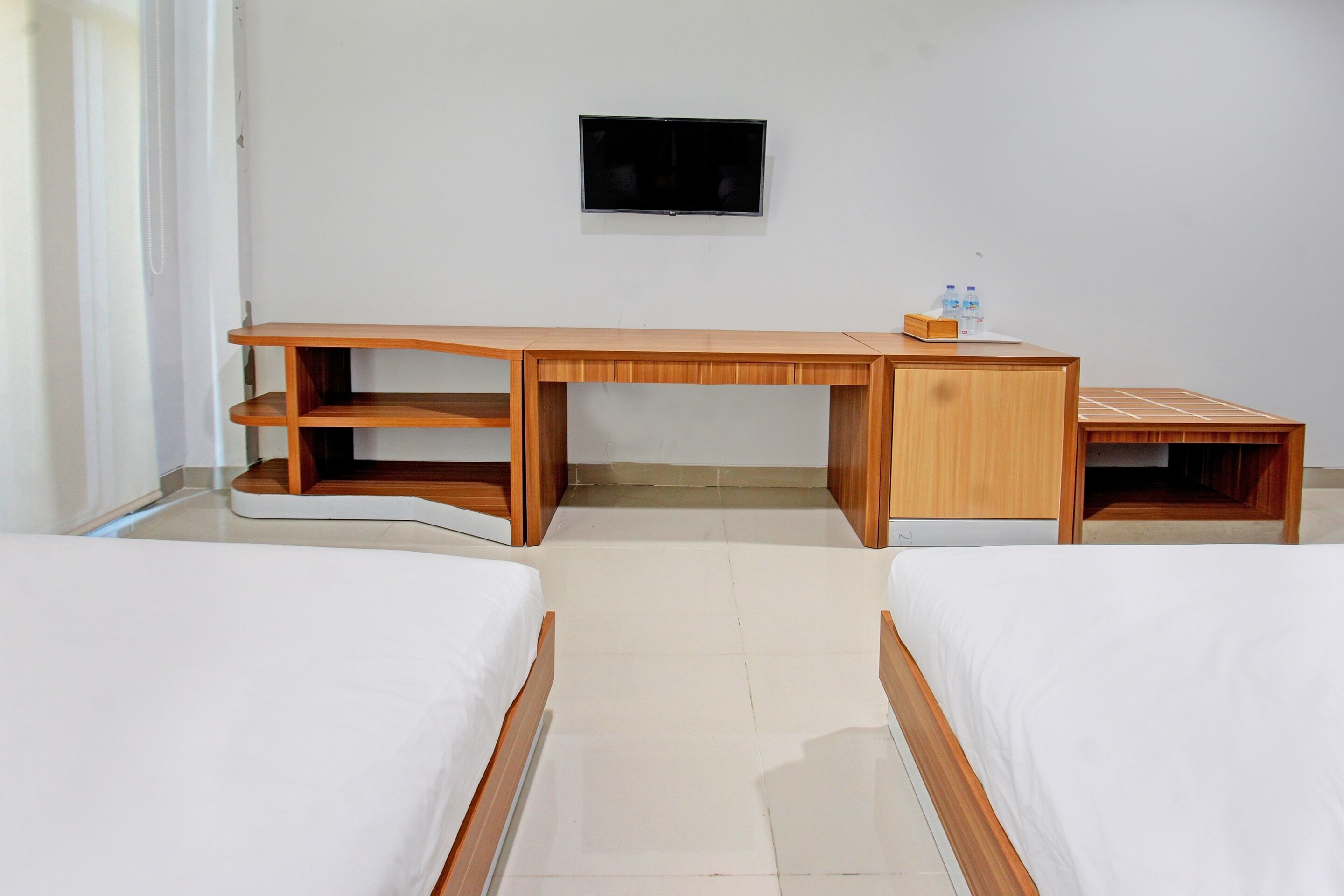 Ein Zimmer mit einem Holztisch, zwei Betten, einem Flachbildfernseher und einem kleinen Regal mit Wasserflaschen.