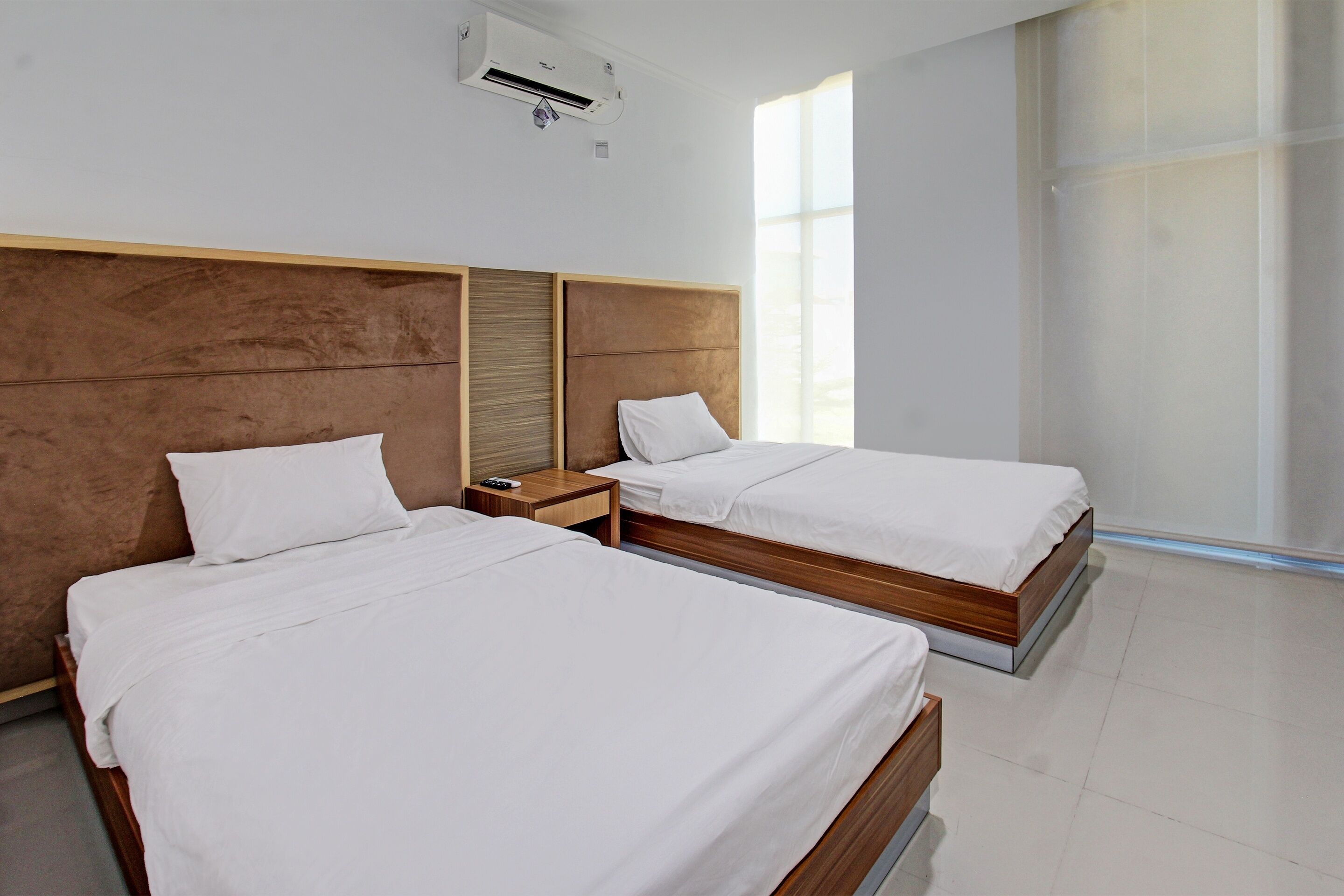 A hotel room with two beds, a wooden headboard, a nightstand, and a window providing natural light.