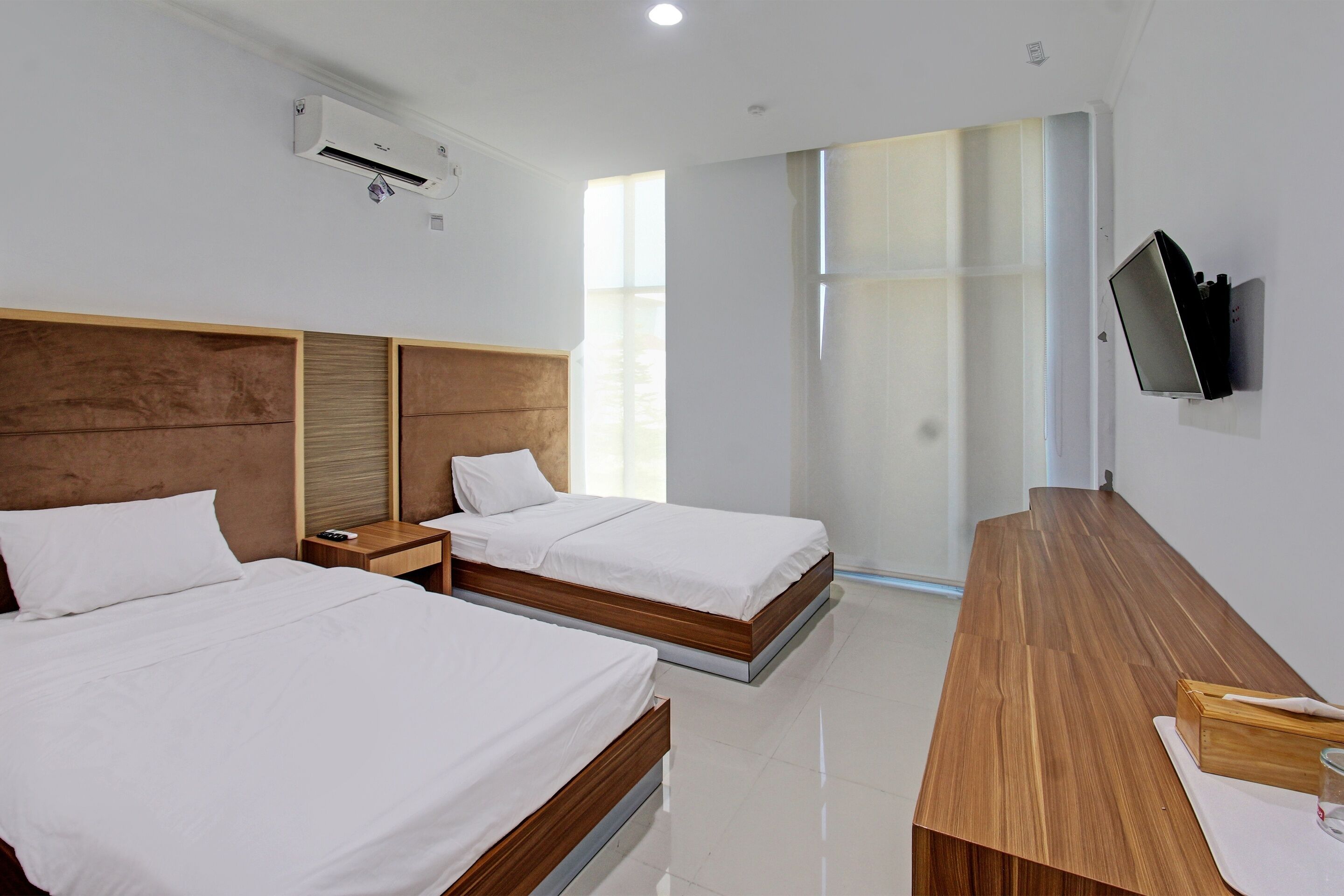 A hotel room with two beds, a wooden desk, a flat-screen TV mounted on the wall, and an air conditioning unit.