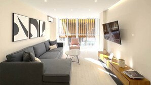 A modern living room with a grey sofa, a round coffee table, a TV mounted on the wall, and a large window with blinds.