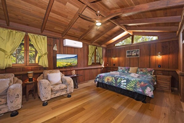 Une chambre confortable, aménagée dans le style d’un chalet, avec des murs en bois, un grand lit recouvert d’une courtepointe à motifs floraux, une télévision et une fenêtre avec des rideaux.