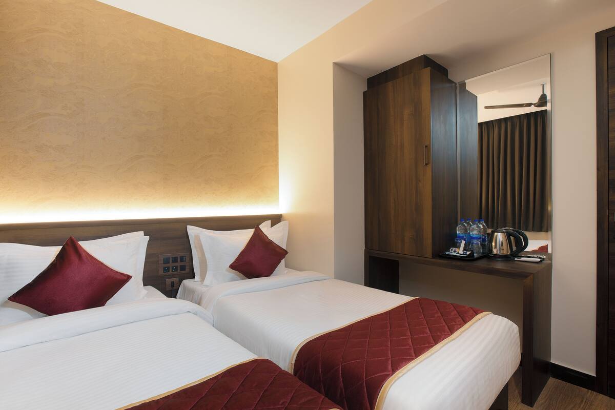 A hotel room with two beds, a wooden wardrobe, a desk with a mirror, and a nightstand with a kettle and bottles.
