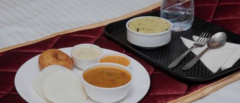 A tray with a plate of food including rice, a soup, and a pastry, along with a bottle of Aquafina water and cutlery on a bed with a red bedspread.