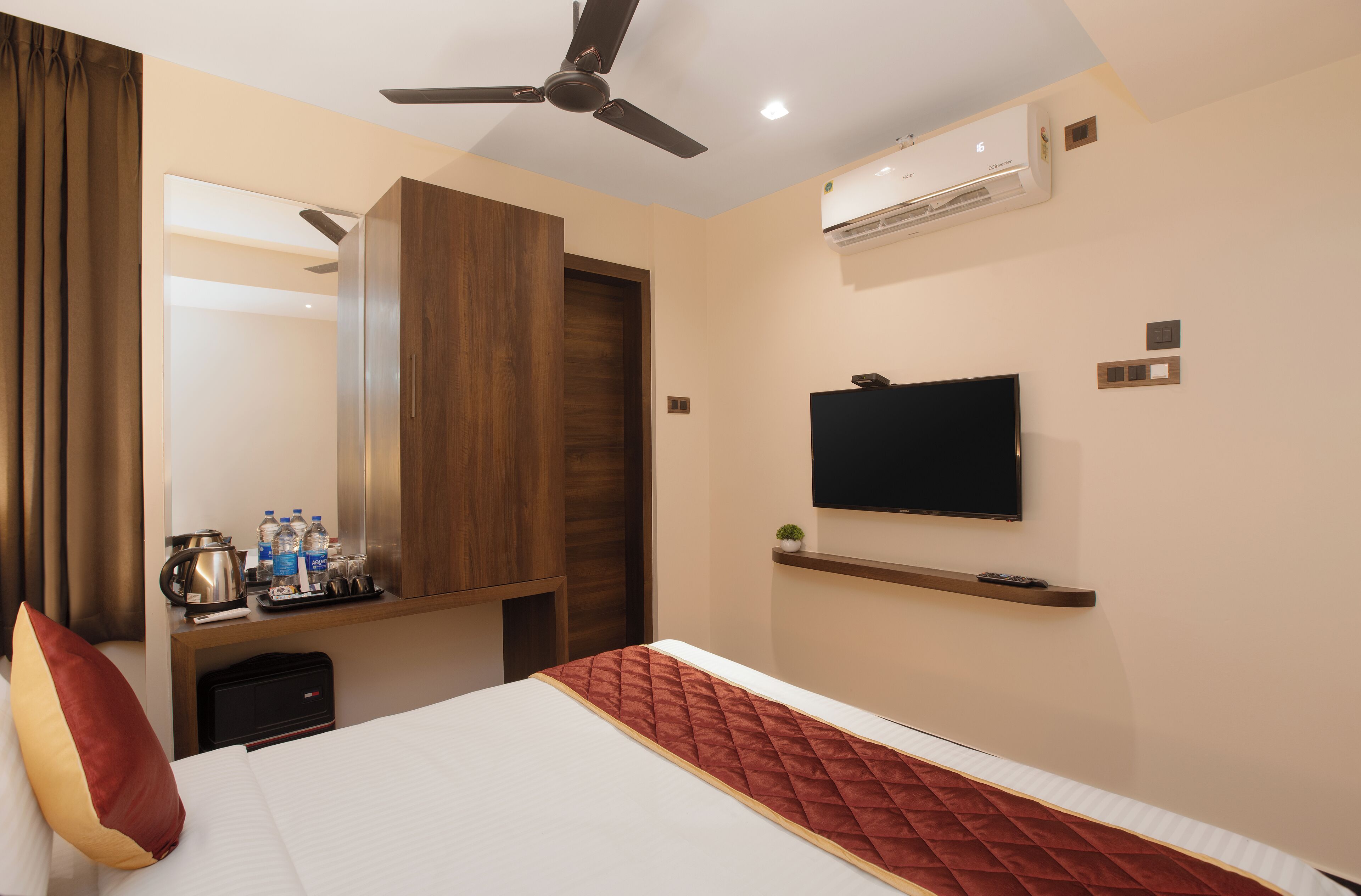 Habitación de hotel con cama, televisor montado en la pared, ventilador de techo, aire acondicionado y un pequeño estante con artículos de aseo.