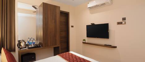 Habitación de hotel con cama, televisor montado en la pared, ventilador de techo, aire acondicionado y un pequeño estante con artículos de aseo.
