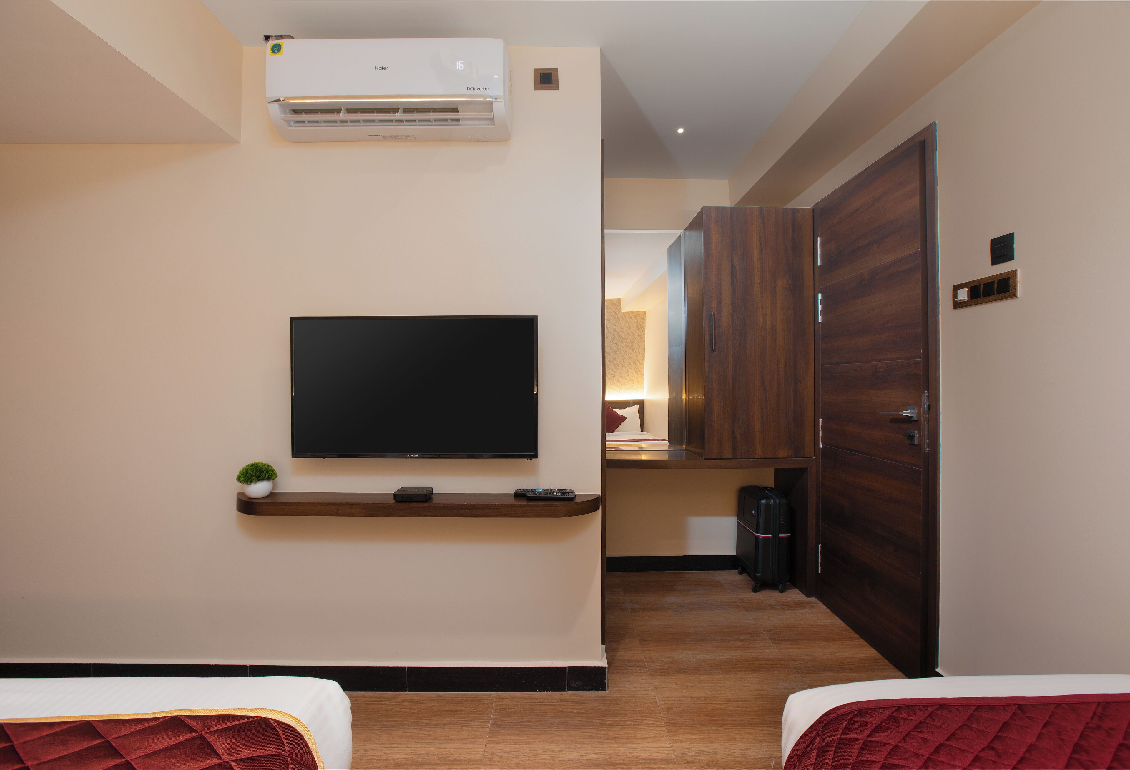 A hotel room with a flat-screen TV mounted on the wall, a small plant on a shelf below, and a wooden wardrobe.