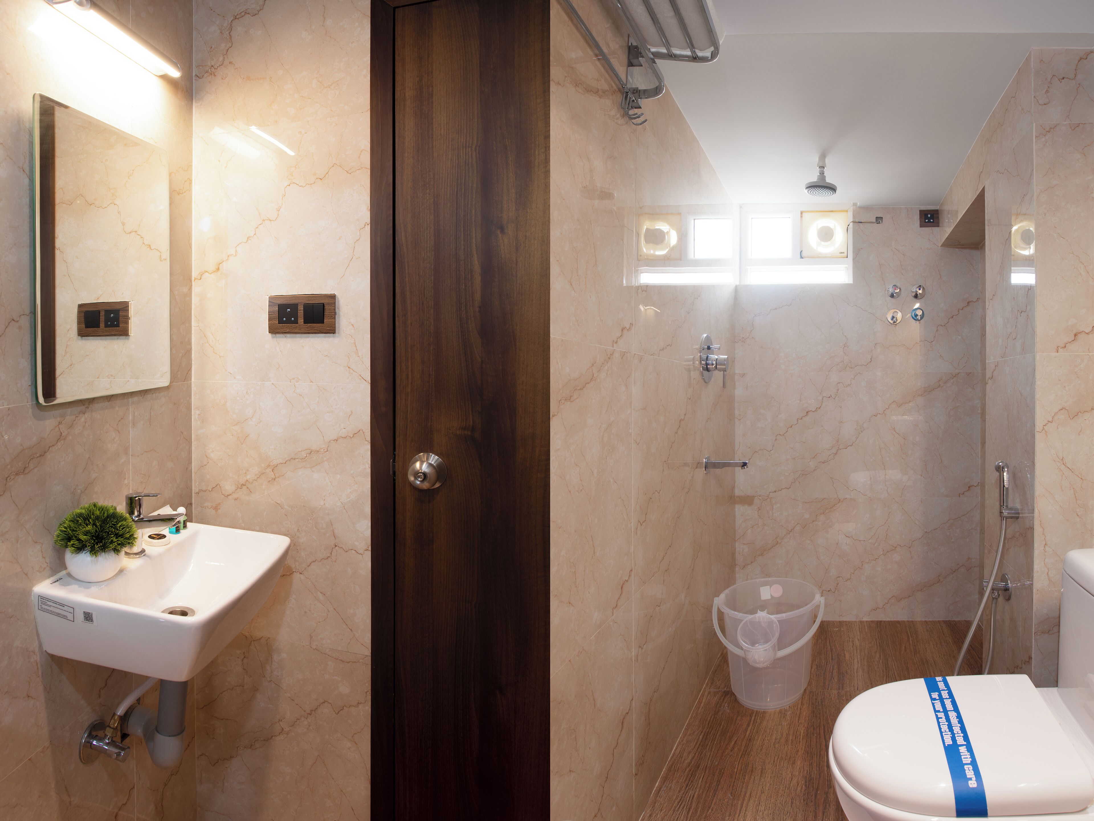 A modern bathroom with a marble sink, a wooden door, a shower with a glass enclosure, and a toilet with a blue safety strap.