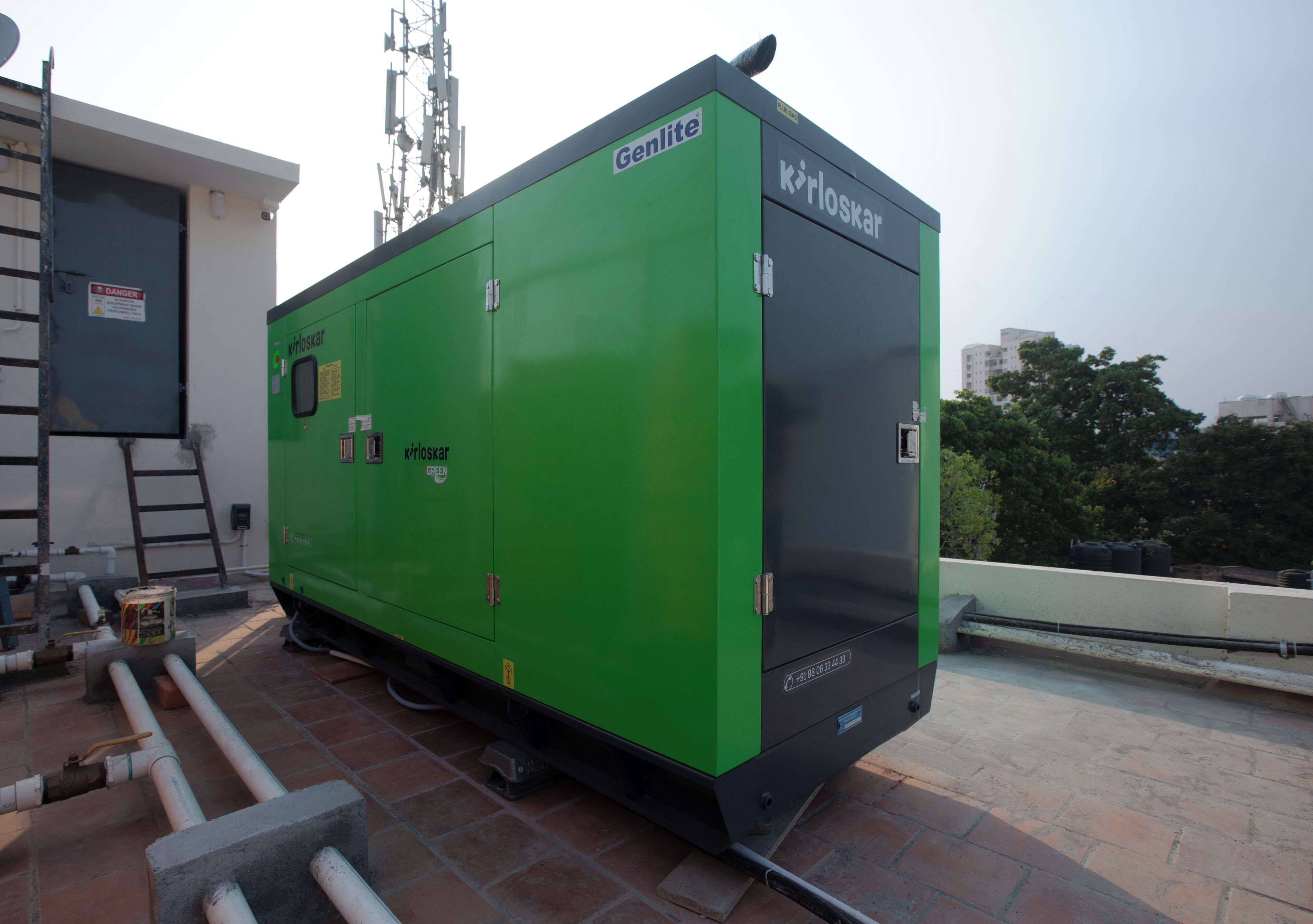A green industrial generator with the brand 'Genlite' and 'kirloskar' on it, situated on a rooftop with visible pipes and a ladder.