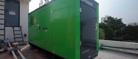 A green industrial generator with the brand 'Genlite' and 'kirloskar' on it, situated on a rooftop with visible pipes and a ladder.