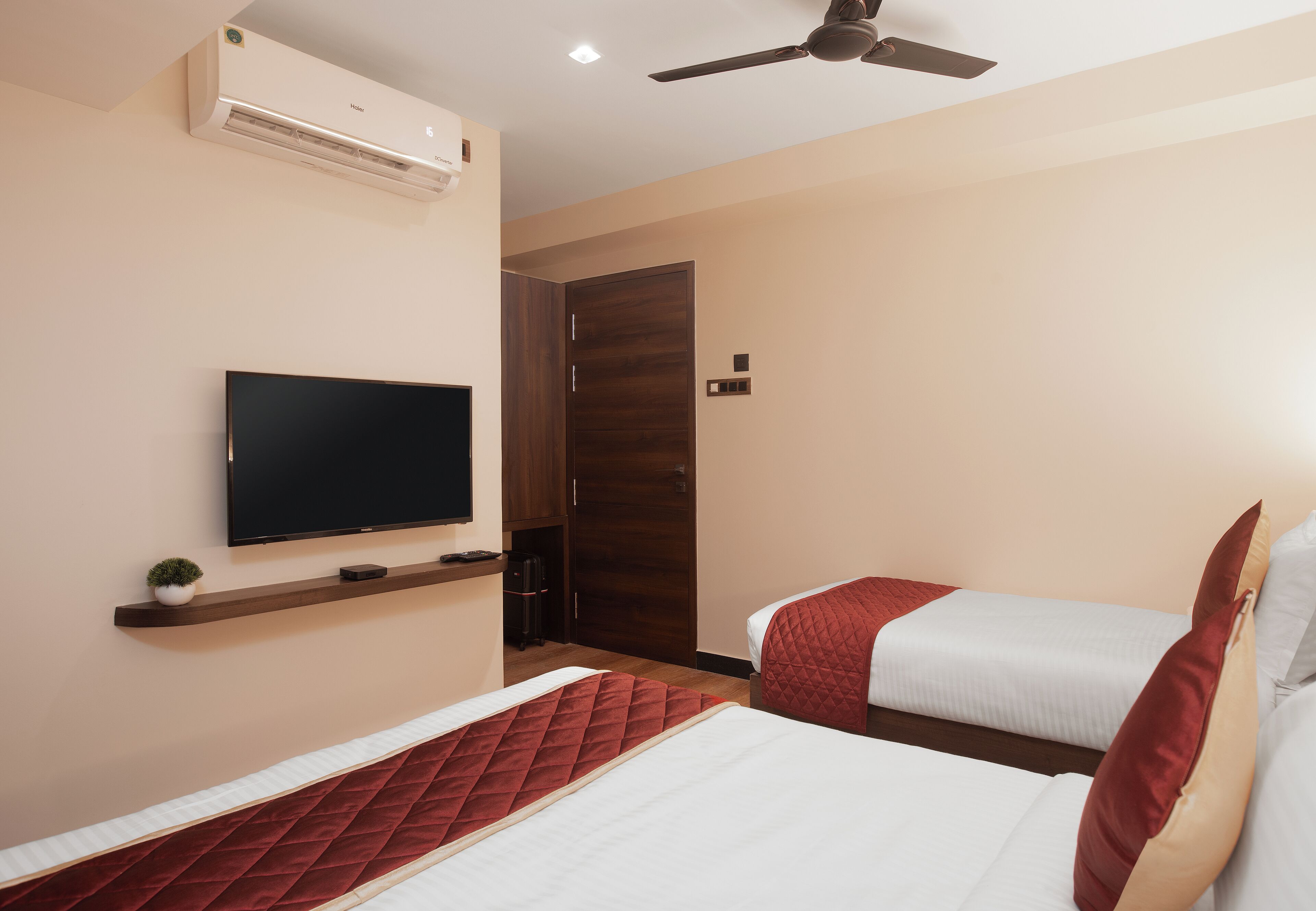 A hotel room with a TV mounted on the wall, a ceiling fan, and two beds with red and white bedding.