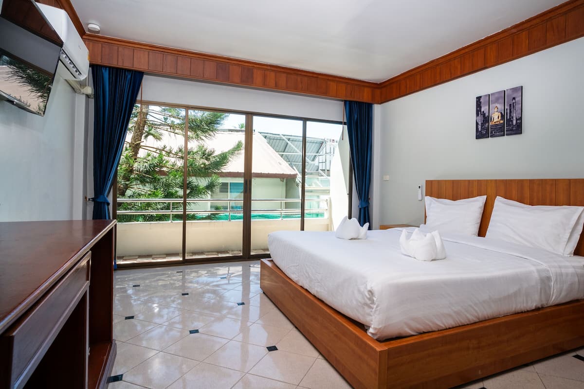 A hotel room with a large bed, wooden headboard, a desk, and a view of a building and trees through the window.