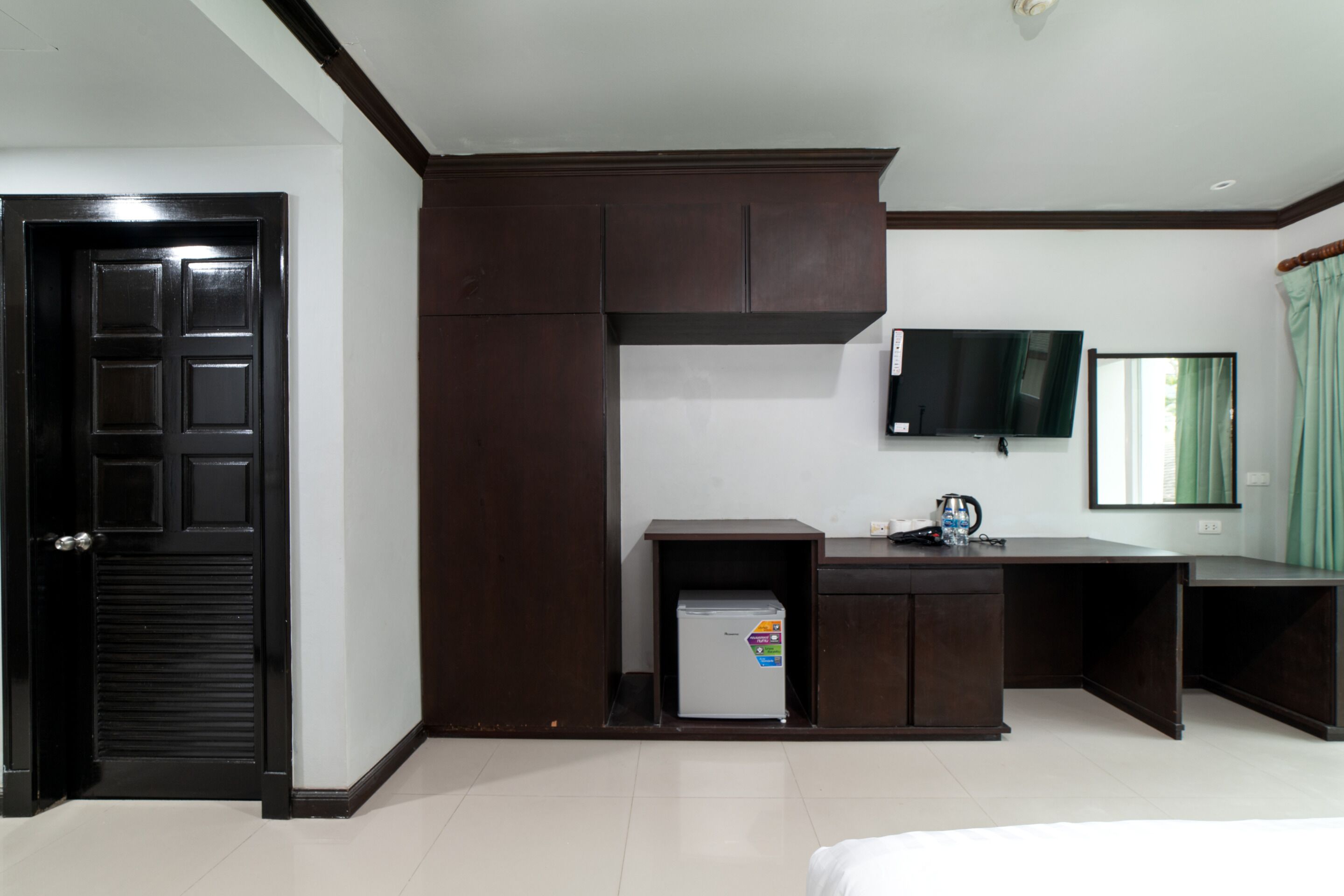 A hotel room with a dark wooden wardrobe, a small refrigerator, a desk, a television, and a bed.