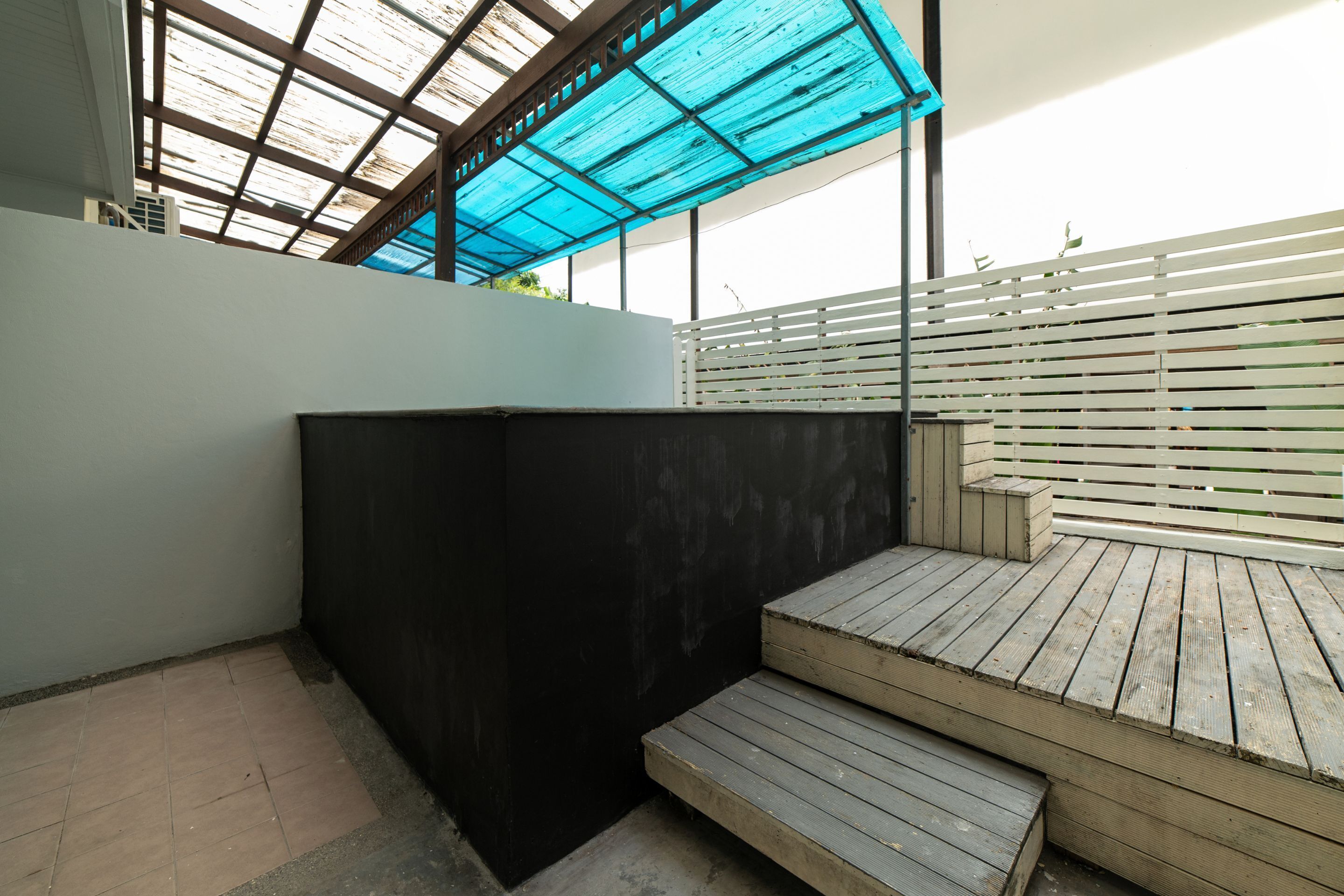Un espacio exterior moderno con una terraza de madera, un muro negro y una estructura de techo azulada.