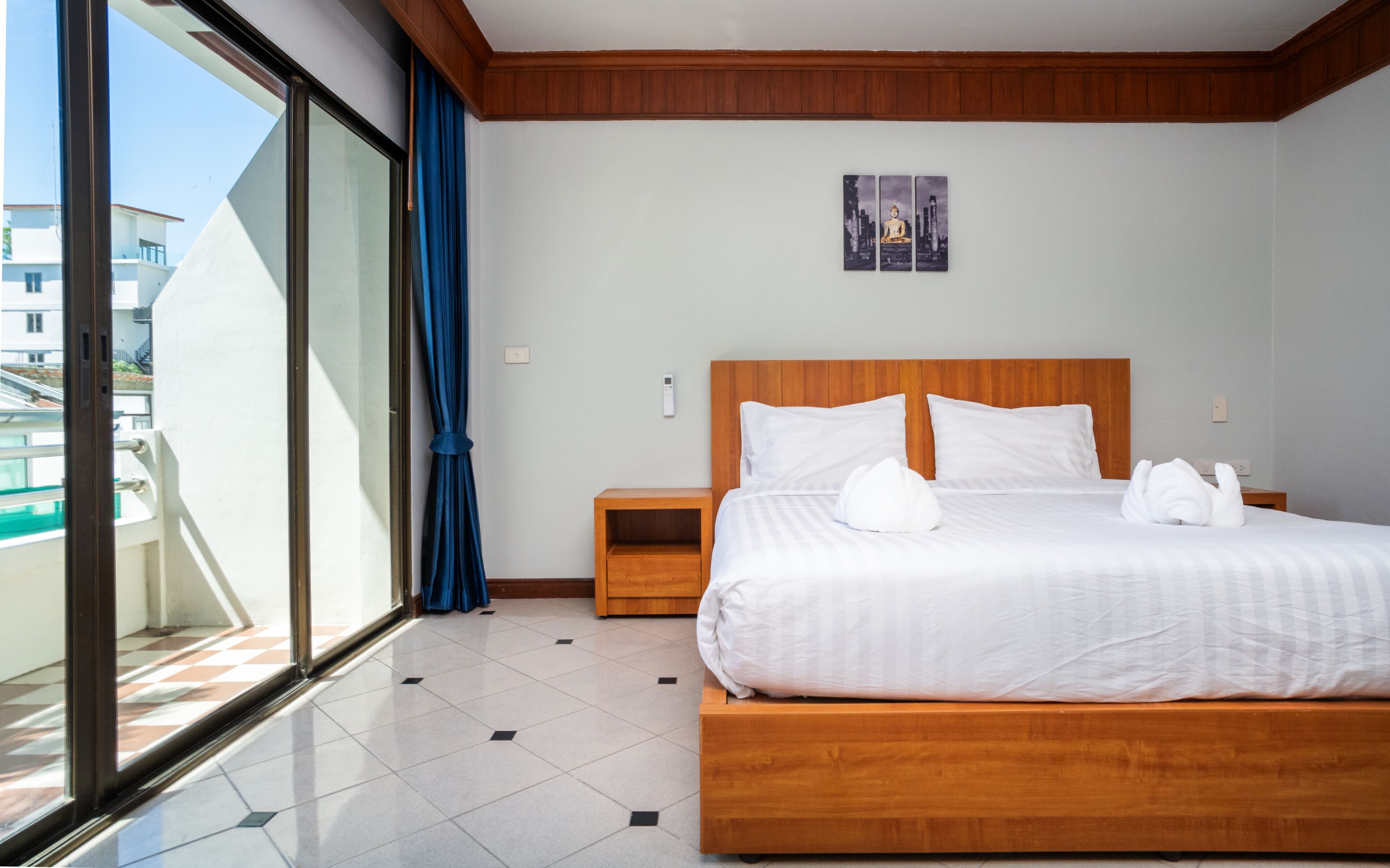 Une chambre d’hôtel avec un grand lit, une tête de lit en bois, une table de chevet, une œuvre d’art accrochée au mur et une vue sur les bâtiments par une grande fenêtre.