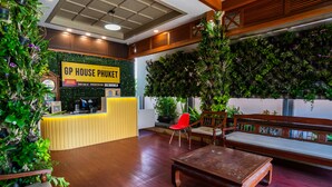 Een receptiegedeelte met een houten bureau, een rode stoel en een bank. Er staan potplanten en een bord met de tekst GP House Phuket.