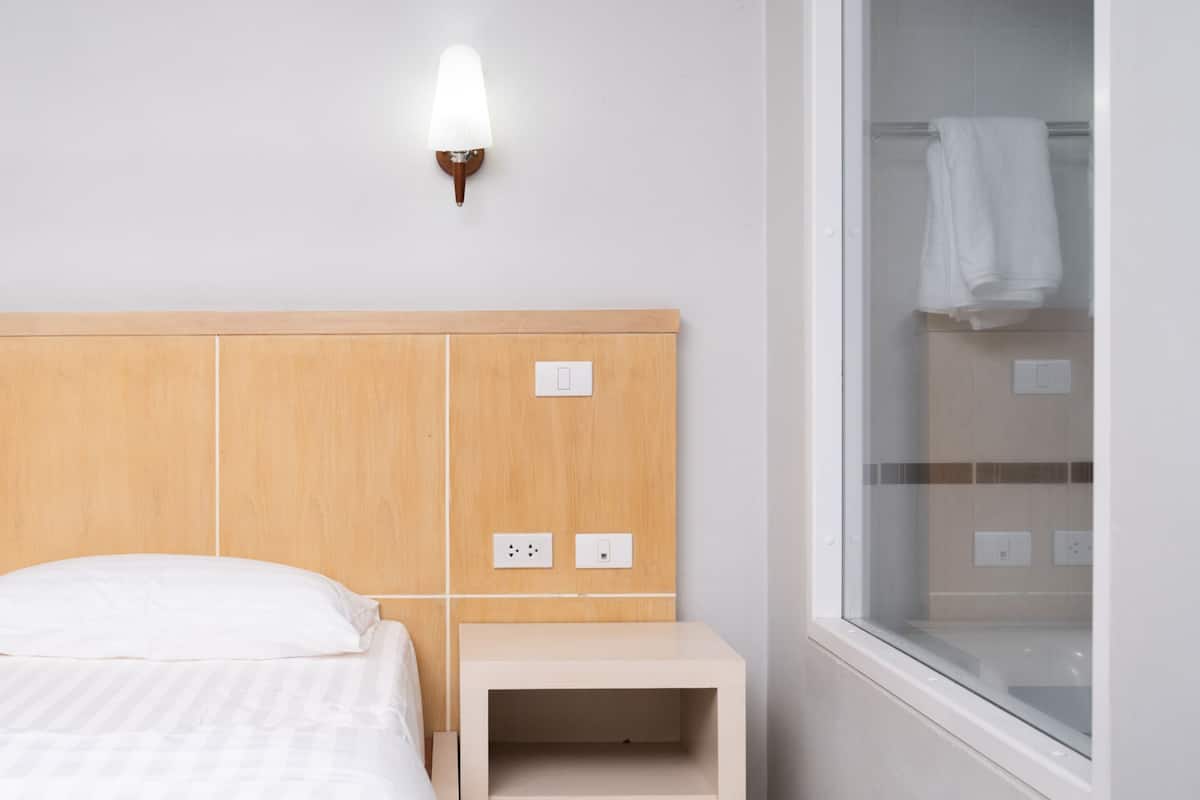 A hotel room with a wooden headboard, a bedside table, a wall-mounted light, and a window with a towel hanging on the inside.