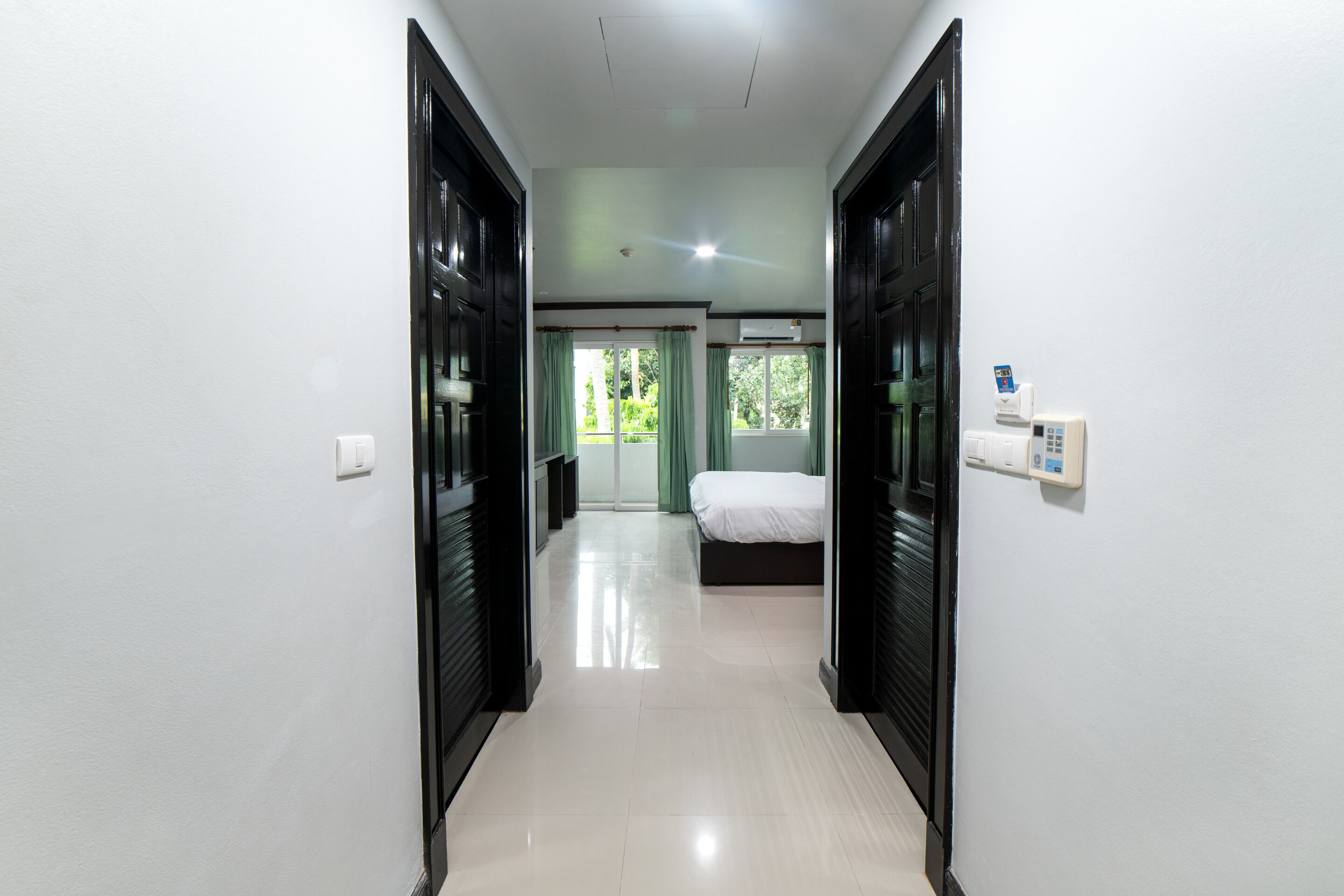 A hotel corridor with a bed, a window with curtains, and a thermostat on the wall.