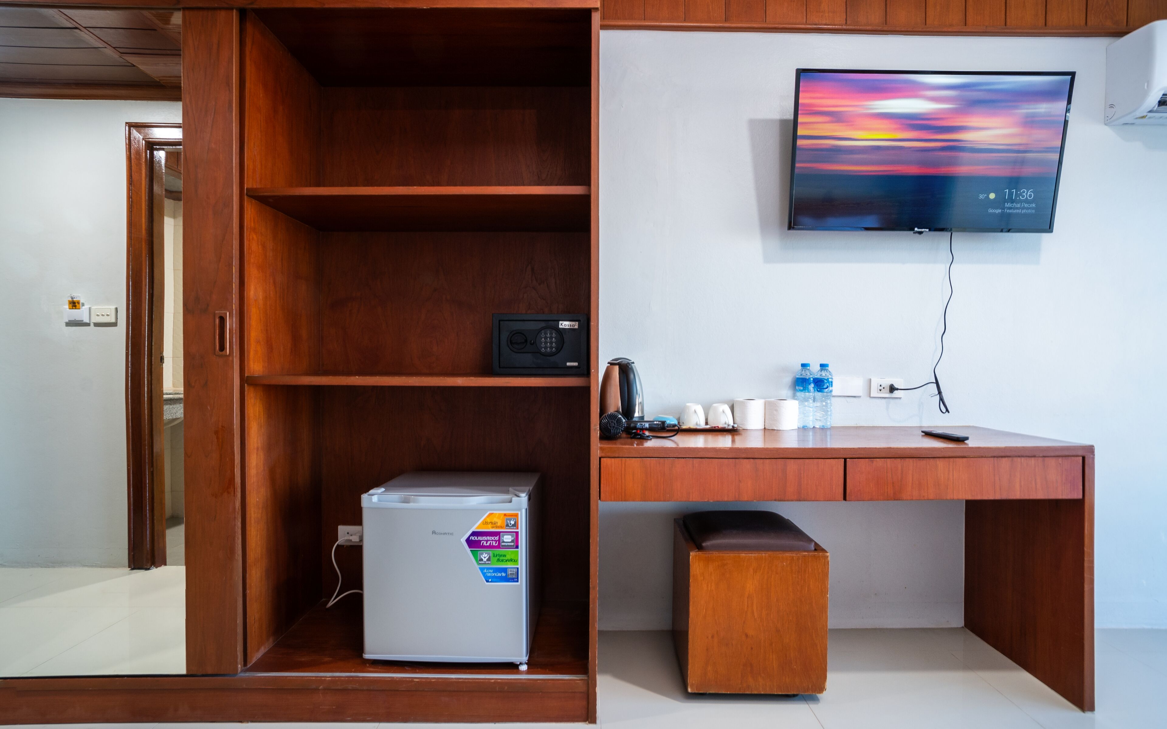 A modern office space with a wooden desk, a refrigerator, a coffee maker, and a television mounted on the wall.