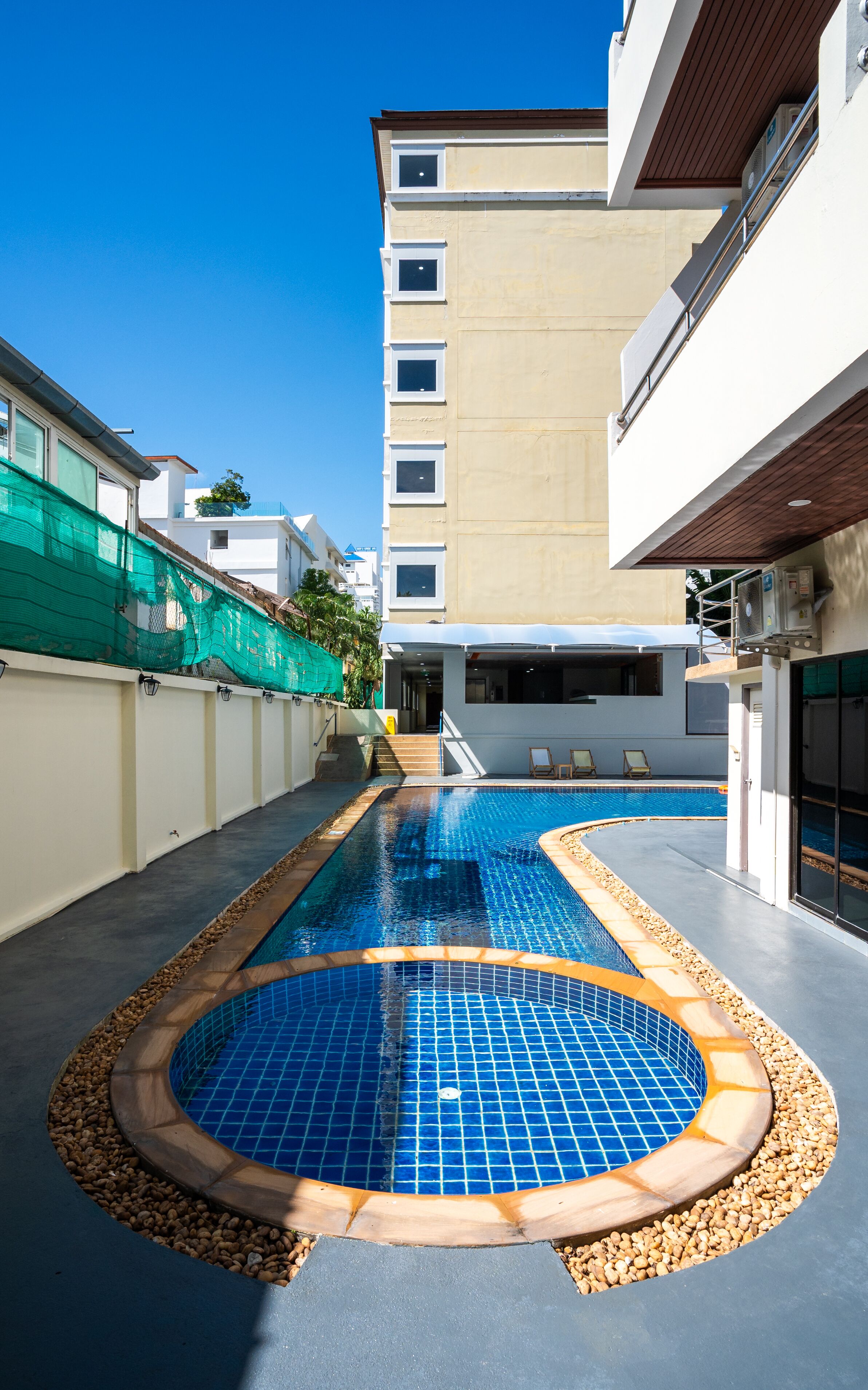 Una piscina con solárium de baldosas, una zona cubierta con sillas y un edificio con varias ventanas y balcones.