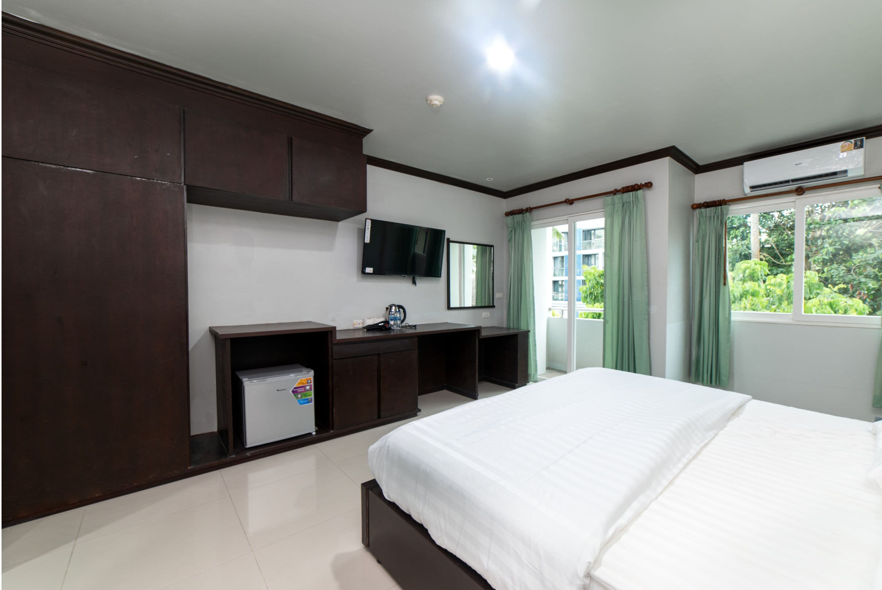 A hotel room with a large bed, a TV mounted on the wall, a small refrigerator, and a window with curtains.