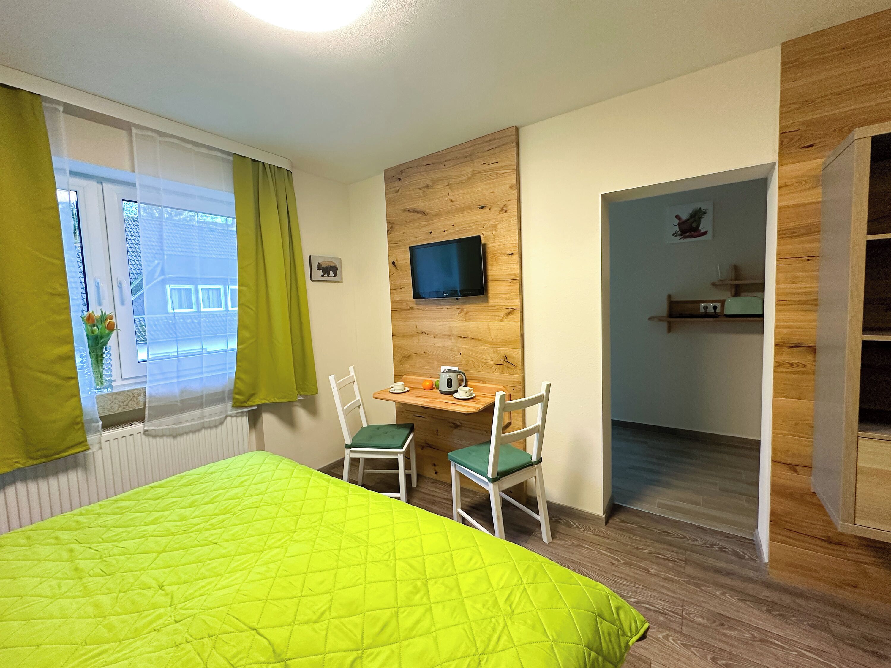 A hotel room with a bed, a desk with a chair, a television mounted on the wall, and a view of the outside through a window with curtains.