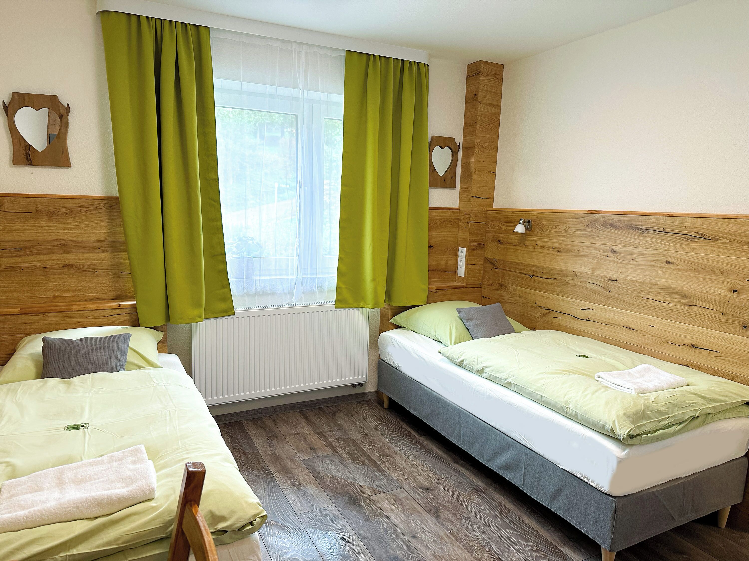 A room with two beds, a window with green curtains, wooden headboard, and a radiator.