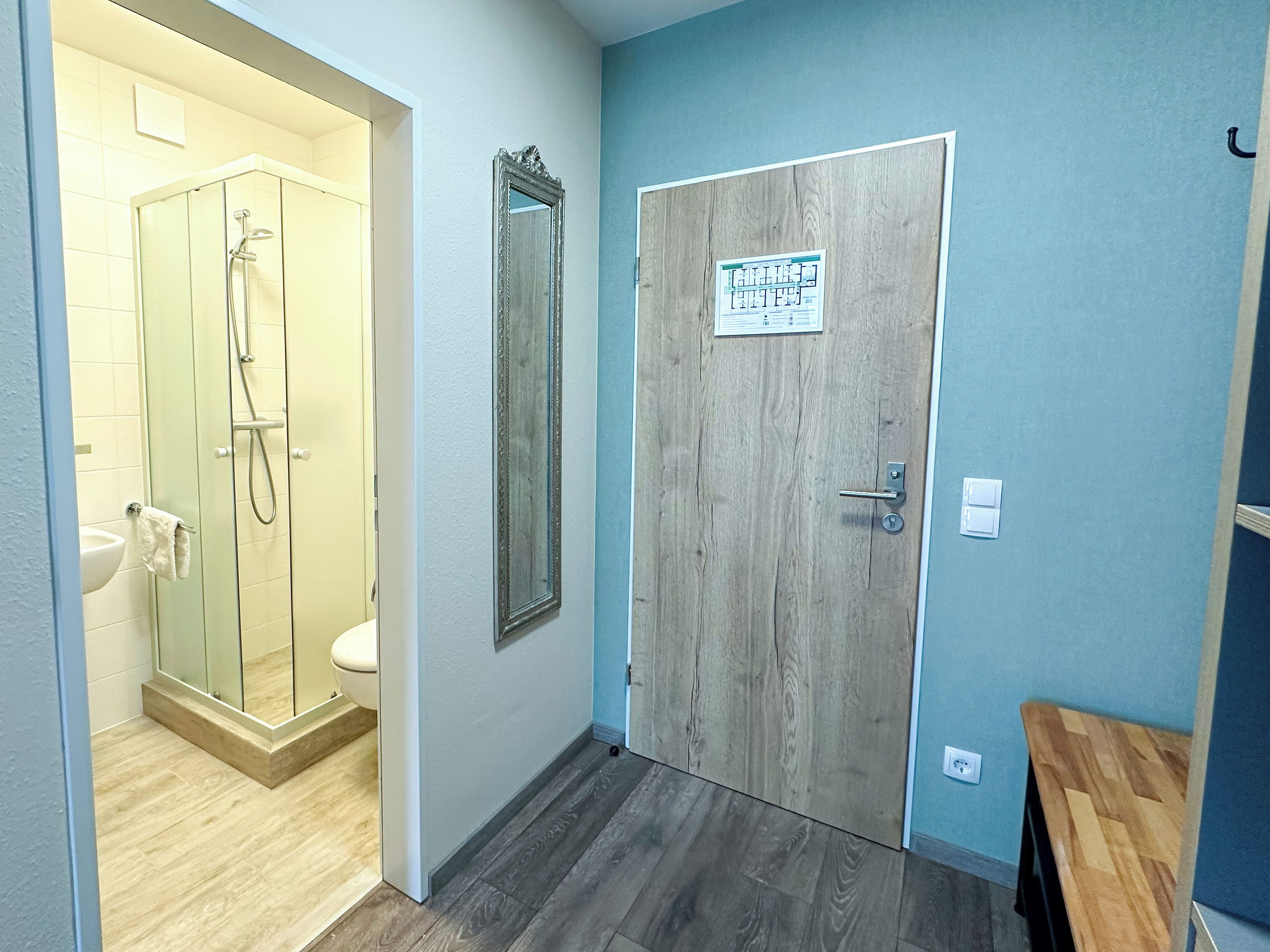 A bathroom with a glass shower enclosure, a wooden door with a sign, and a wooden floor.