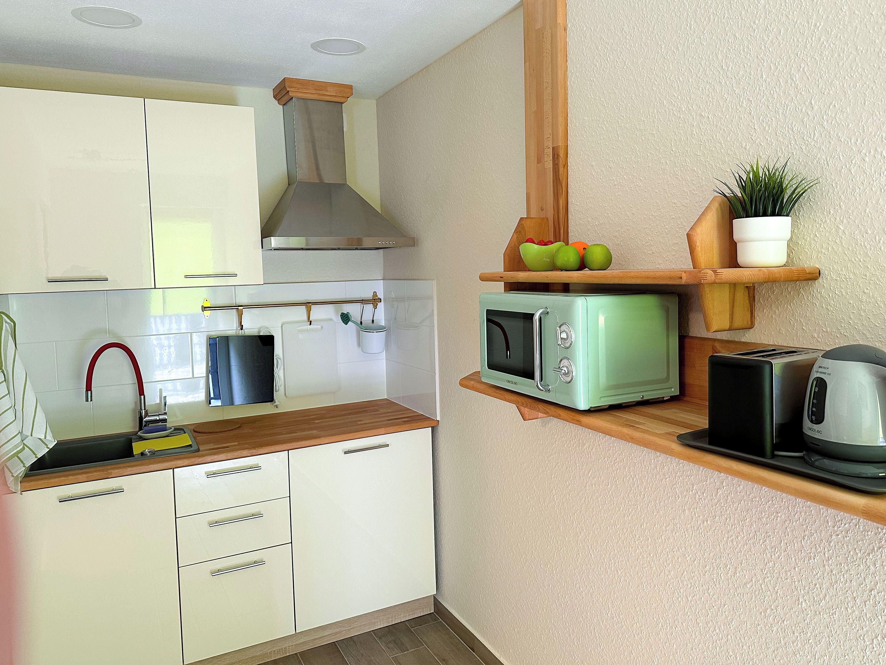Una cocina compacta con armarios blancos, microondas, tostadora y estante de madera con frutas y una planta.