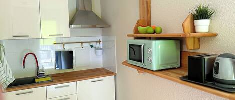 Una cocina compacta con armarios blancos, microondas, tostadora y estante de madera con frutas y una planta.