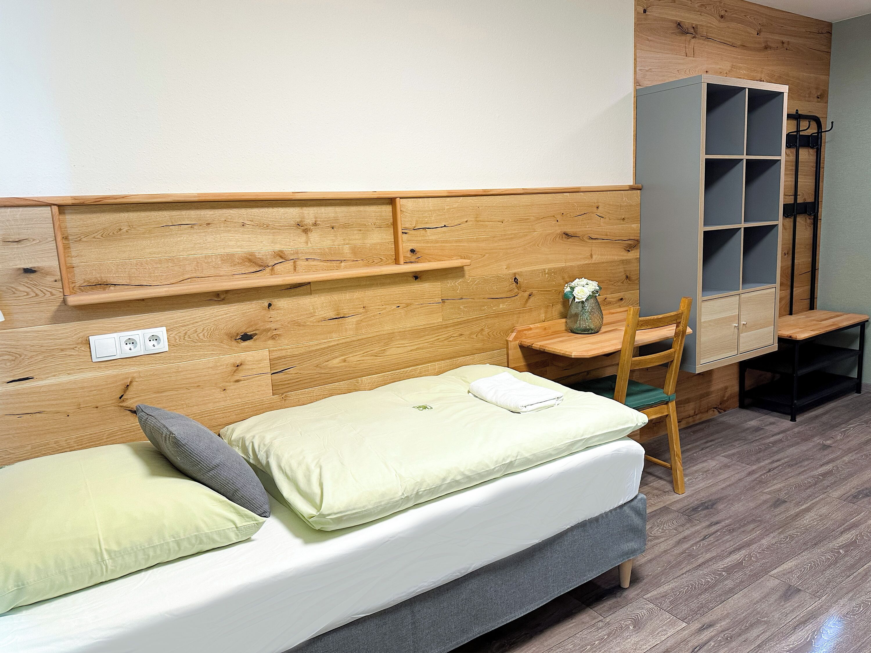 A small, cozy room with a wooden headboard, a single bed with pillows, a small table with a vase, and a built-in shelving unit.