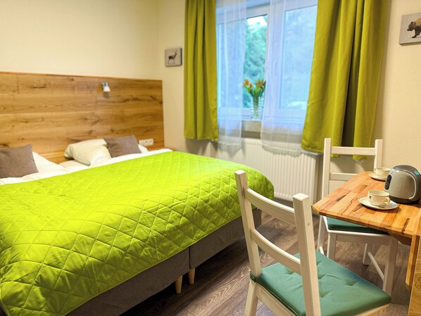 Hotelkamer met een bed, een houten hoofdeinde, een raam met gordijnen, een tafel met waterkoker en een stoel met kussen.