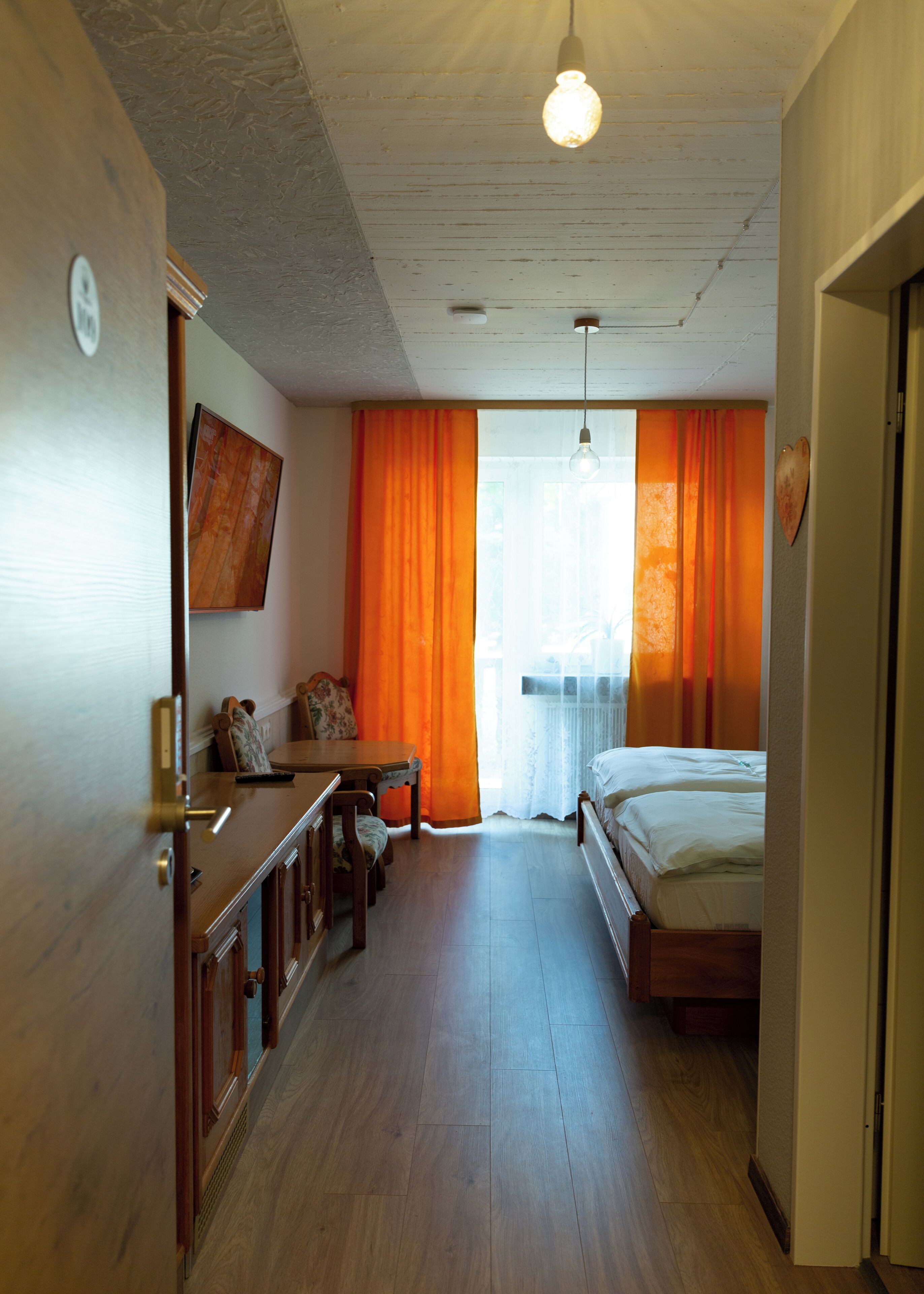 Ein Hotelzimmer mit Holzboden, einem Bett, einem Schreibtisch mit Stuhl und einem an der Wand befestigten Fernseher.