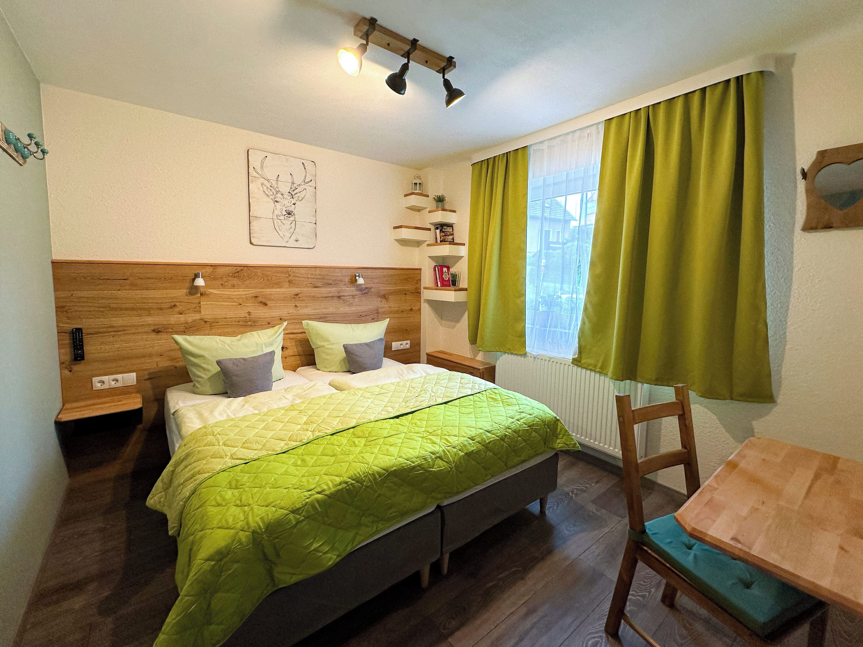 A bedroom with a wooden headboard, a bed with green bedding, a bedside table, a nightstand, a small wooden table with a chair, a window with green curtains, and a wall-mounted shelf with books.