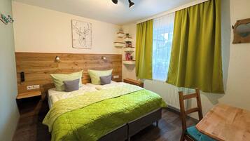 A bedroom with a wooden headboard, a bed with green bedding, a bedside table, a nightstand, a small wooden table with a chair, a window with green curtains, and a wall-mounted shelf with books.