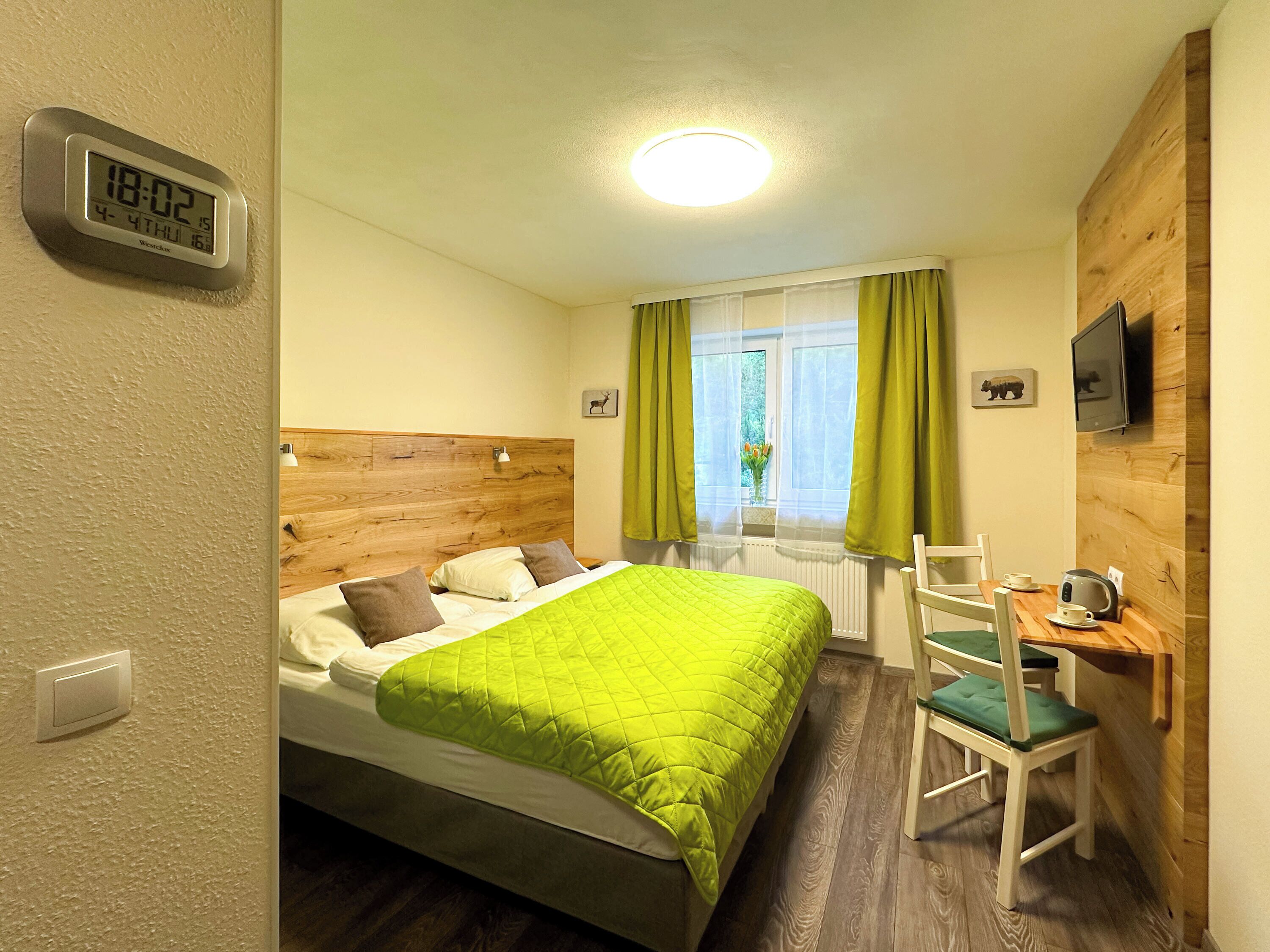 A hotel room with a bed, a chair, a table with a kettle, a TV, and a window with curtains.