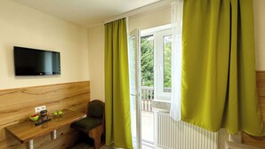 A small room with a wooden bench, a TV mounted on the wall, and a window with green curtains. - Hotel Waldberg (Stolberg)