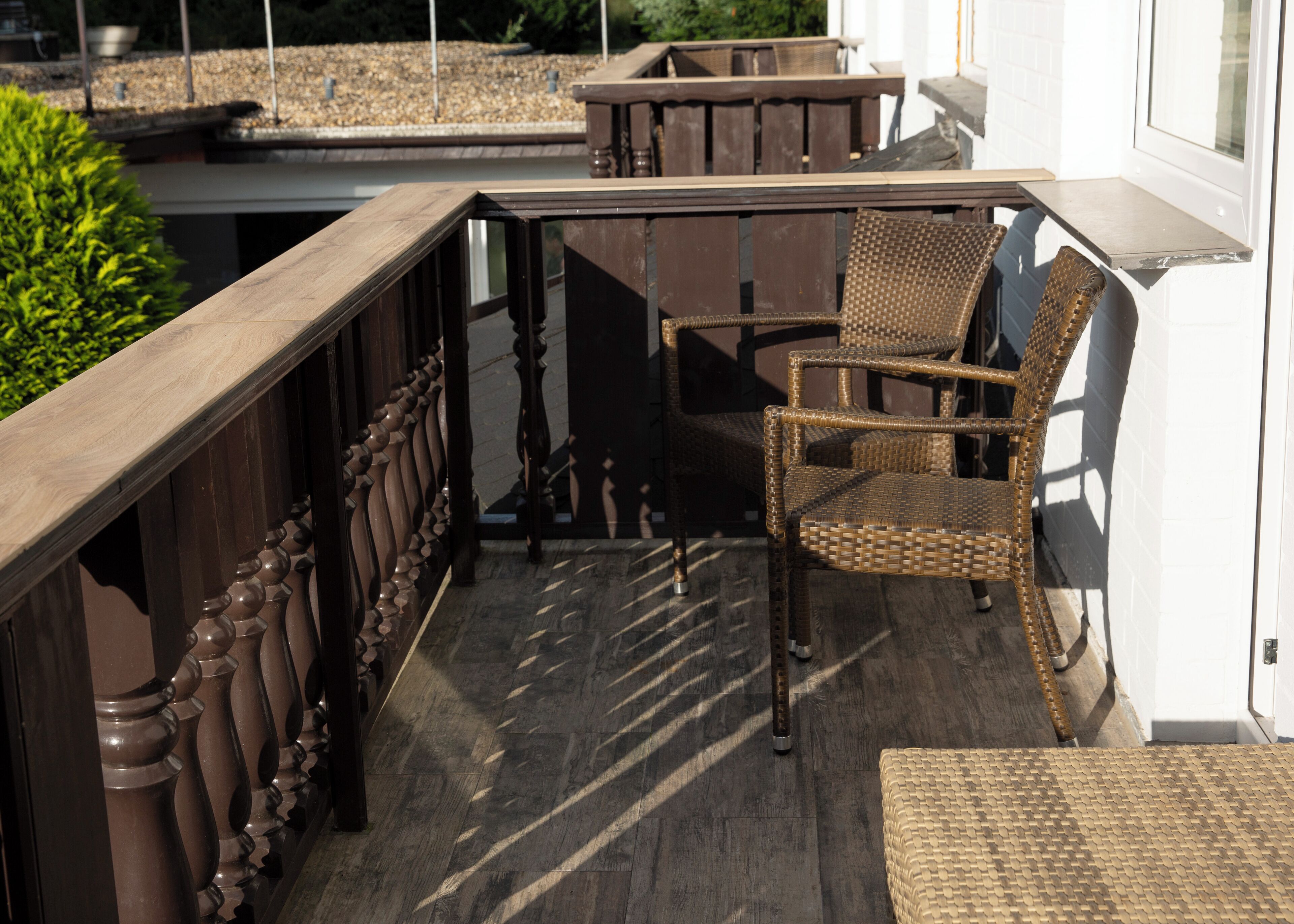 A balcony with a wicker chair and a small table, wooden balustrade, and a view of greenery.