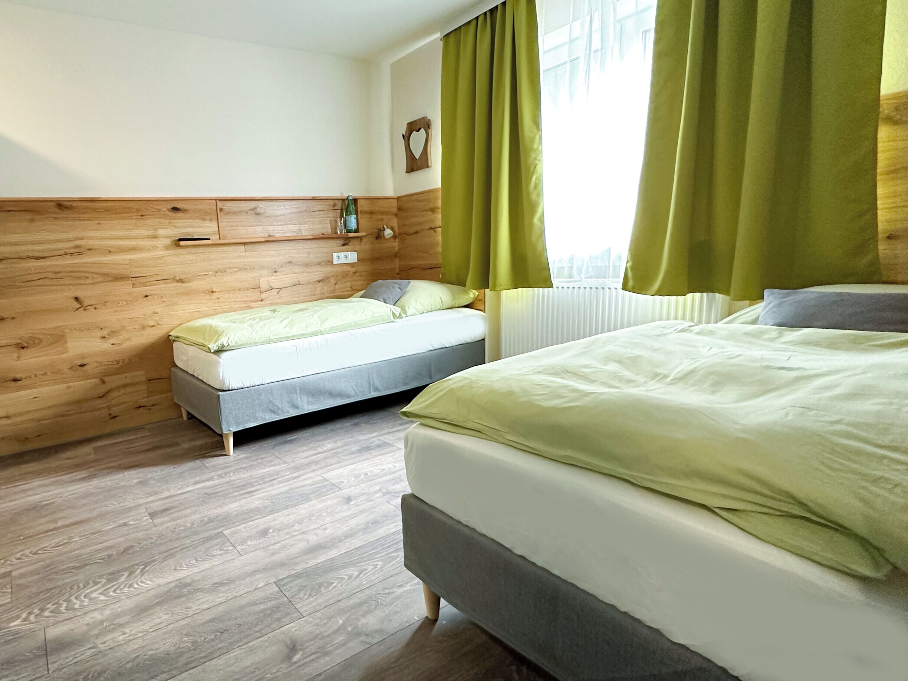 A hotel room with a wooden wall, a bed, a smaller bed, a nightstand, a window with curtains, and a radiator.