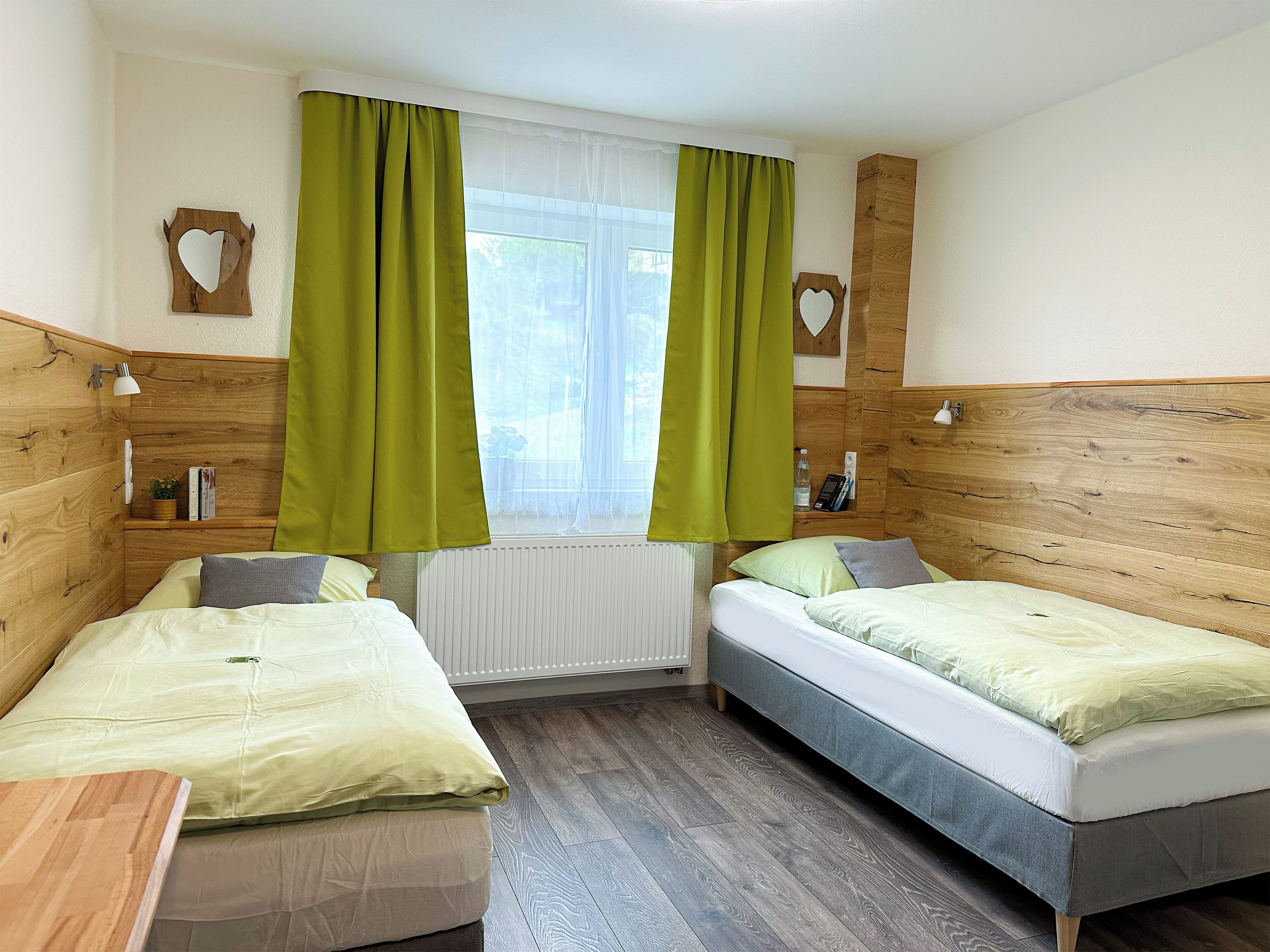 A room with two single beds, a window with sheer curtains, a radiator, and wooden headboards.