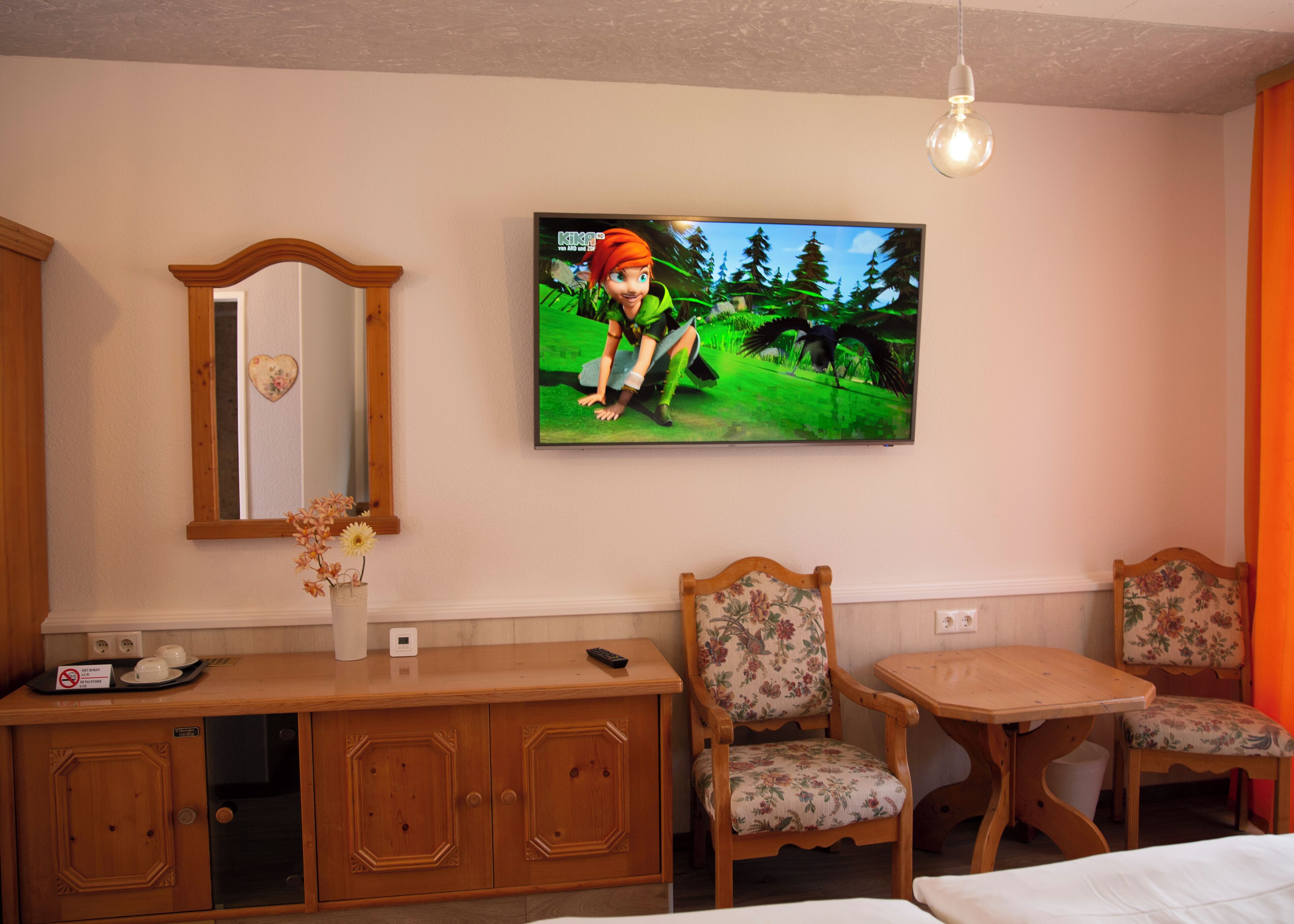 A hotel room with a wooden dresser, a mirror, a television displaying a cartoon character, two floral chairs, and a small table.