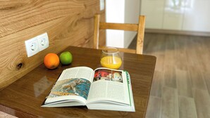 A wooden table with a book, two fruits, and a glass of orange juice in a room with a wooden bench and a kitchen in the background. - Hotel Waldberg (Stolberg)