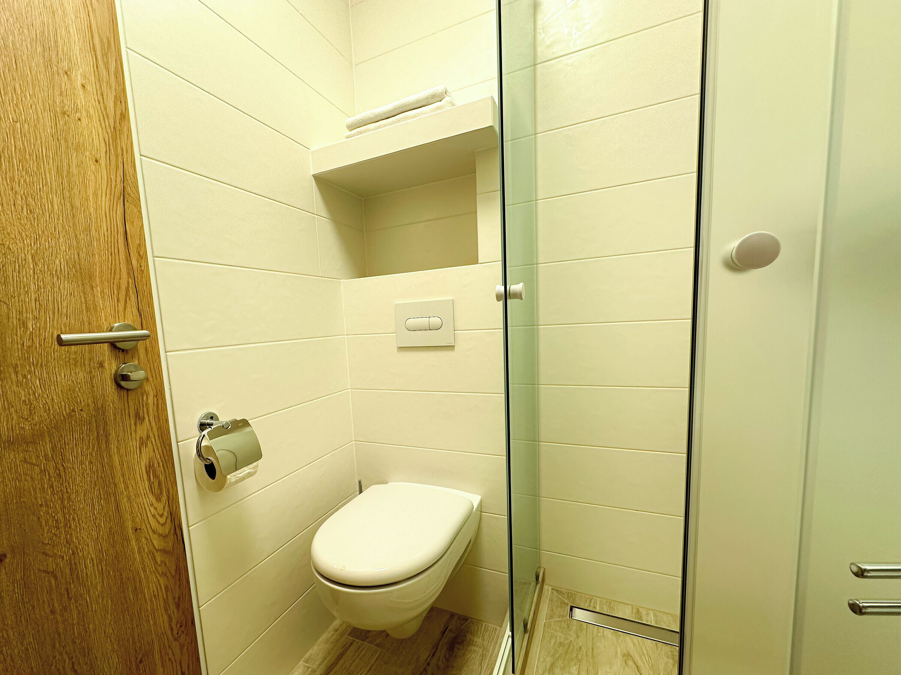 A small, modern bathroom with a toilet, a glass shower enclosure, and a wooden door.