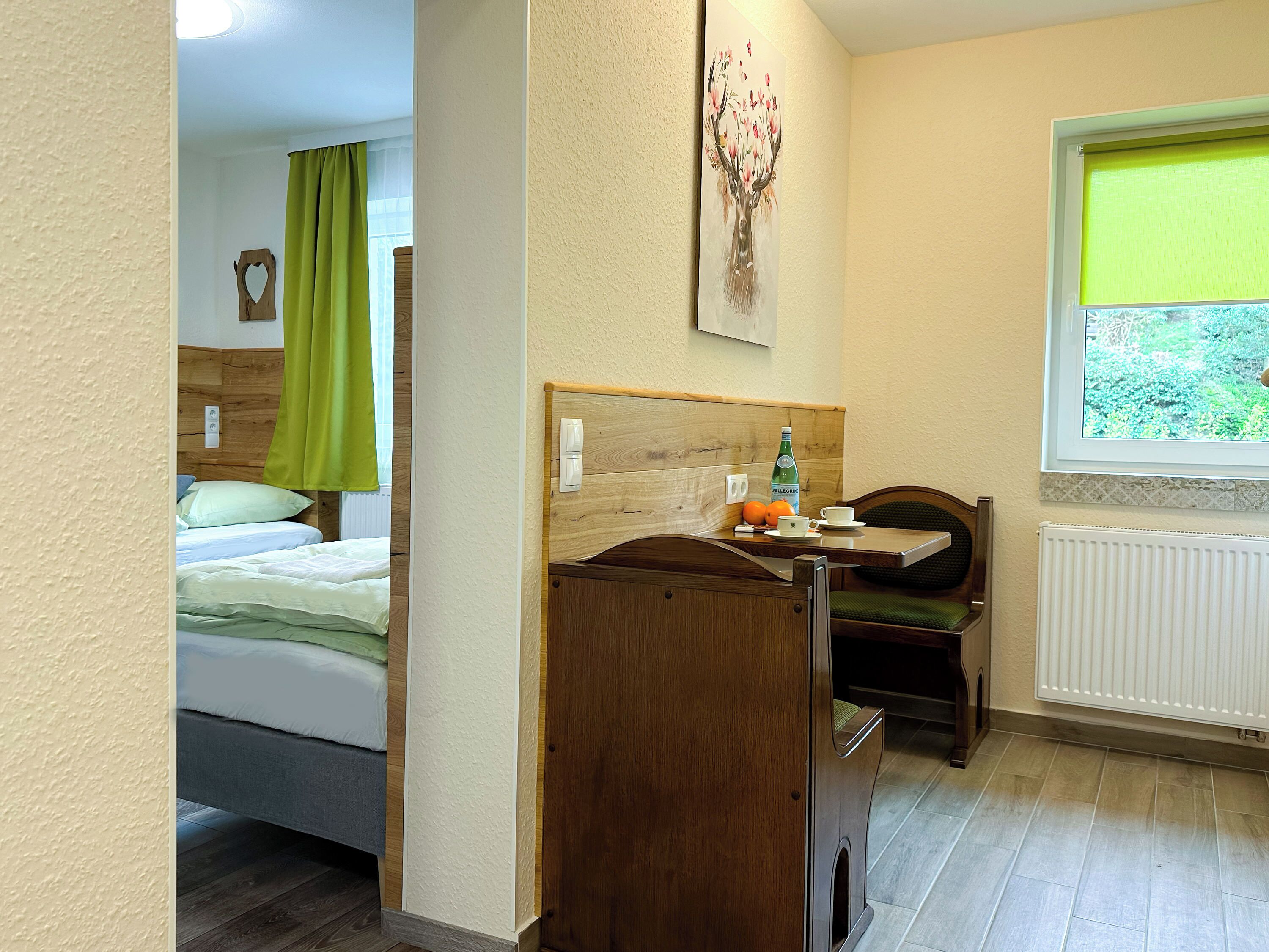 A hotel room with a bed, a wooden desk with a bottle, a cup, and oranges, a radiator, and a window with a view of greenery.